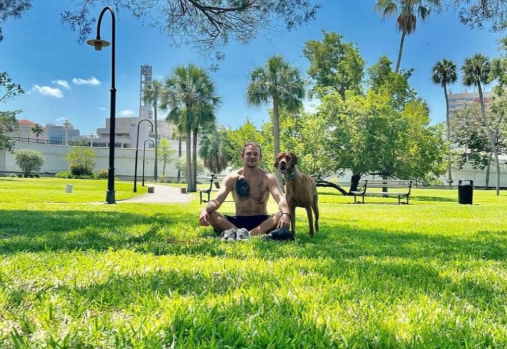 Man sitting in the grass with a brown dog standing at his side