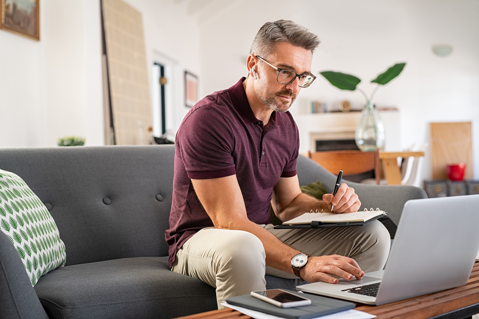 man sitting on couch writing in notepad while on laptop